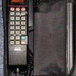 Unzipped black bag phone, open to view interior contents, including a brick-sized phone receiver and curly cord.