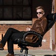 Person wearing brown leather jacket and sunglasses, sitting on black office chair outside