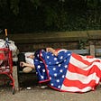 A houseless man sleeps on a bench next to a shopping cart full of his belongings, draped in an American flag blanket.