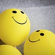 Three yellow balloons on a grey background. Two have visible black smiley faces.