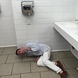 A young man in a seersucker sport jacket, white chinos, and poorly matched dark brown lace-ups, flips off the camera as he lays on a bathroom floor in a drunken stupor.