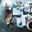 Color photo of a bluish desk of pens, X-mas cards, festive rubber stamps, a teapot and cup of tea.