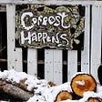 Hand painted sign on a rustic fence saying “Compost happens” above a pile of wood.
