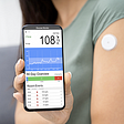A woman wearing a continuous glucose monitor on her upper arm holds out a phone showing her blood glucose data