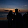 Darkened photograph of a pair of lovers with a background of sunset in Bali and a number of out-of-focus people in the frame