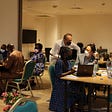 A view of a room with participants from a Hackathon organized by the UN country team in Guinea. Three square wooden tables can be seen arranged in the room, with 2 to 4 participants per table seated on light blue chair with gold metal frames. RC Vincent Martin is standing next to two participants at a table.