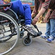 Picture of a person in a wheelchair, and another person standing, who appears to be assisting the first. Their faces are not in the frame. Courtesy of the author.