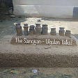 A traditional cooking setup from a tribal village with “The Sangyan: Ulgulan Talks!” written over it on the smaller platform and below the earthen chimney. The setup has eight small earthen chimneys over a small platform that settles over a larger platform all made up of soil and other natural ingredients and are brown in colour.