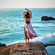 woman in pink flowing dress standing on boulder looking out over turquoise ocean with arms over her head