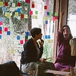 Three people having a discussion in front of a window with lots of post-it notes on it.