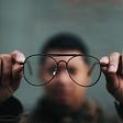 Blurry photo of man holding up a pair of glasses to denote looking at things with a different lens or perspective.