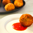 A fried arancini ball on a white plate with a dollop of red marinara sauce in the center.