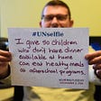 A man holding an #Unselfie sign. He handwrote on it: “I give so children who don’t have dinner available at home can eat healthy meals at afterschool programs.” The date is GivingTuesday, Dec 3, 2019