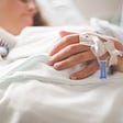 Close up on the hand of a woman asleep in a hospital bed. There’s an IV in the back of her hand.