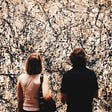 A man and woman stand with their backs to the camera, looking at at abstract piece of art on a wall.