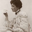 A lady from the Victorian era in black and white photo. We see her left profile. She is seated. Hair is up in bun/chignon with pearl earring visible in left ear. Right hand is holding a small teacup in the air with the forearm barely touching table. Left hand is laying in her lap. She is wearing a dress with buttons at the cuffs covering wrists; with buttons, collar, and tie going up the neck and covering it. The only part of her exposed is head and hands.