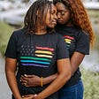 Two feminine-presenting people with dark skin are embracing, one person standing behind the other with their hand wrapped under the other’s arm and resting on the other’s stomach. Both are wearing black shirts with rainbows in the shape of U.S. Flag stripes. Their faces are nuzzling close together, the shorter person in the front looking back and the taller person’s pierced nose tilting downward. Their eyes are closed and they seem at peace in their intimacy.