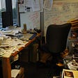 A home office room that is very messy. Papers, CDs and other things litter the desk and the shelves around. A white board with several sheets of paper sticking to it can be seen in the background.