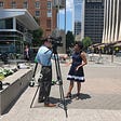 Kristen is wearing a blue dress with a white stripe and she’s standing next to a reporter in a blue shirt and green kakhis and their camera on the Fayetteville Street Mall in Downtown Raleigh, NC in June of 2019