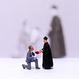 A man in a grey suit holding a bouquet of red flowers is down on one knee holding the hand of a man in a long black robe and hat who is standing.