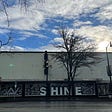 Old storefront, windows painted with white garland and large marquee sign with the word shine. Parting clouds and sun above.