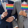 Gay male couple in wedding suites with arms interlocked hold little gay pride flags.