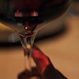 Extreme close-up of a right hand offering a stemmed glass of deep red wine against a gold-hued, soft-focus background.