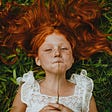 A redheaded girl lying on the grass holding a plant in front of her face.
