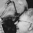 A monochrome image of an elderly couple with their foreheads touching and smiles on their faces.