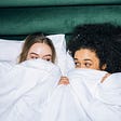 Two women lay in bed next to each other, hiding under the blankets. They look at each other flirtatiously. #lesbians #sapphic #flirting #sex #bed #lesbiansex #kilvinmethod