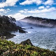 Waves breaking on a rocky coast under a partly cloudy sky.