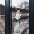 A photo of a woman wearing a mask looking out the window from her home.