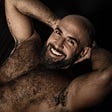 Handsome middle aged man with arms behind his head and no shirt on, smiling widely. Very hairy.