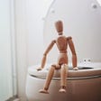 A wooden artist’s model is posed sitting on a toilet