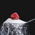 Close-up of a spoon (extending from the right) full of spilling sugar, with a raspberry on top. Black background.