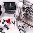A table consisting of a Mac running a 3D modeling program , a 3D pritner , plus 3D toys and other mechanical tools.