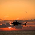 Helicopter flying over water, with a sunset hidden behind clouds in the background. The sky is very orange.