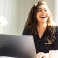 A woman sitting at a desk with a laptop and a mug in front of her and she is smiling broadly — medium.com/@kristin_missk — self-improvement — self-development — rituals — routines — writing tips
