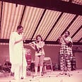 Three people on a stage playing the flute. There is a white woman in middle and two African men in traditional African dress to either side of her.