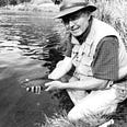 Larry Salata kneels on a river bank with a trout in his hand