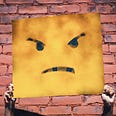 Two hands hold up a square sign with an angry face on it in front of a painted red brick wall.