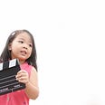A young Asian girl holds a black and white film clapper and looks off to the right of the frame.