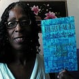 Author Charm Baker (aka Justiss Goode) holds up blue African American Heritage book by author Dennis W. DeLoach