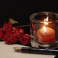 A burning candle within a glass holder, next to a fountain pen and dried roses.