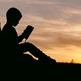 sillouette image of a child meaning against a tree reading a book
