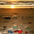 A colour photograph of a sandy beach looking out to sea at the approach of sunset. In the foreground the beach is littered with a significant amount of plastic waste of varying size, shape and colour.