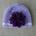 A light purple crochet hat with one strip of white toward the bottom and big, purple flower on the side