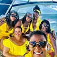 Six black women dressed in yellow swimwear and sunglasses on a boat, smiling.