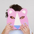 A girl hides behind a paper mask of a pink lion.