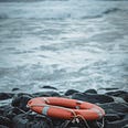 A picture of a round, orange life bouy sitting on the rocks and the waves hit against them. “Like many, I am a survivor. Okay, that sounds so cliche. It’s not and you will learn why more over time.”